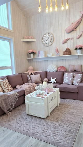 Gemütliches Wohnzimmer mit einem Kamin, einer großen Fensterfront, einer L-förmigen Couch in Rosa, einem Couchtisch, dekorativen Kissen und einem Teppich. Eine Treppe führt nach oben, und es gibt Regale mit Dekorationen.