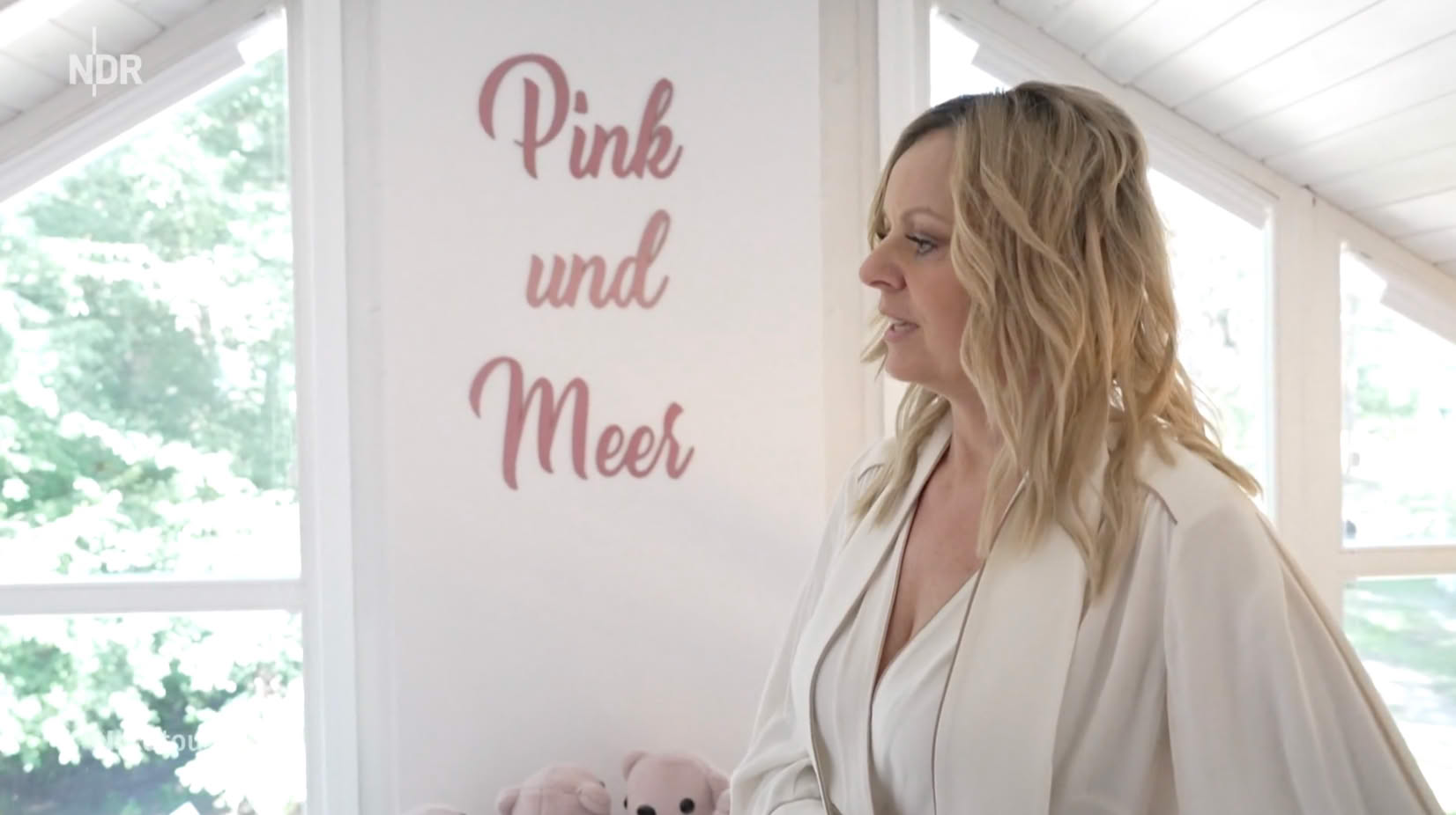 Eine Frau mit langen blonden Haaren steht in einem hellen Raum vor einem Fenster. An der Wand steht in rosa Schrift "Pink und Meer".